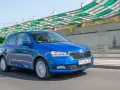 Niebieska Škoda Fabia jedzie po drodze, za nią widać nowoczesną konstrukcję z zielonym zadaszeniem.