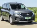 Szary Nissan Townstar jedzie po drodze, otoczony zielonymi polami. Kierowca w okularach prowadzi pojazd.