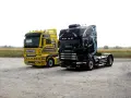 Dwa ciągniki siodłowe: żółty MAN i czarna Scania z grafiką. Różne rodzaje naczep czekają na załadunek.