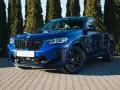 Błyszczący, niebieski suv BMW X4 M Competition zaparkowany przed żywopłotem.
