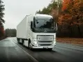Biała ciężarówka Volvo FH12 z naczepą jedzie po mokrej drodze wśród jesiennego lasu.