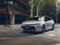 Srebrna Toyota Corolla kombi z czarnymi felgami sunie po miejskiej ulicy, otoczona zielenią i nowoczesną architekturą.