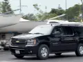 Czarny Chevrolet Suburban jedzie obok przyczep z łodziami, w tle drzewa.
