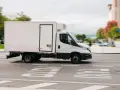 Biała chłodnia, kategoria C1, pędzi ulicą. W tle zamazane drzewa i budynki.