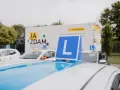 Na dachu samochodu nauki jazdy widoczny znak "L". W tle ciężarówka z napisem "JA ZDAM". Ile jest pytań na kat B?