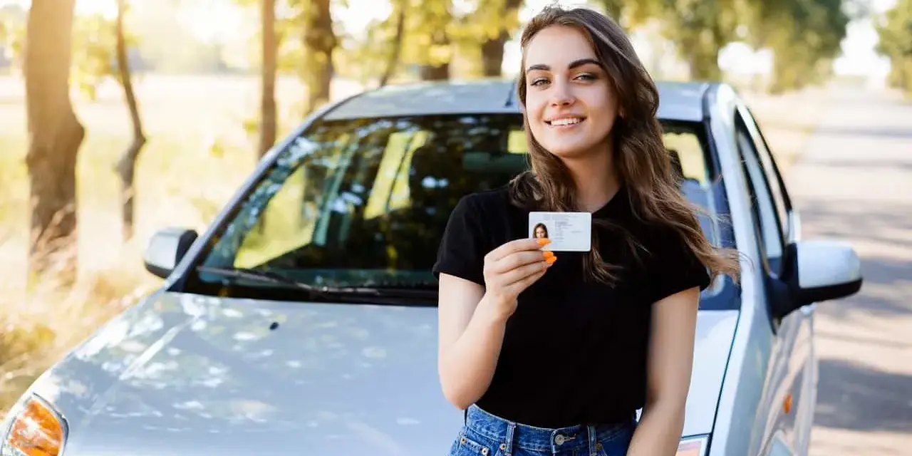 Szczęśliwa młoda kobieta z prawem jazdy od 16 lat stoi obok samochodu na tle drzew.