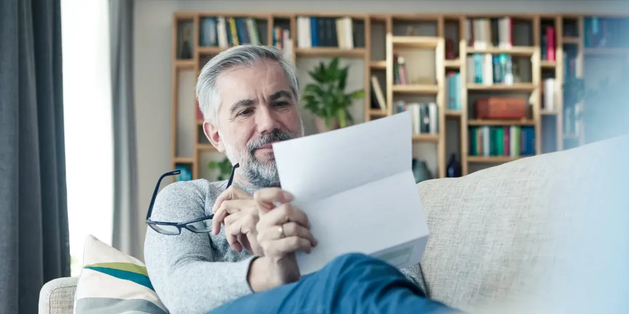 Mężczyzna z siwą brodą czyta dokument, być może wypowiedzenie OC. Na półkach za nim widać książki.