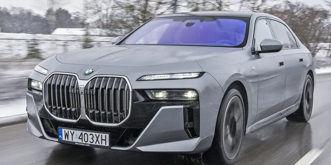 Szary BMW M7 pędzi po mokrej drodze, otoczony zimowym krajobrazem.