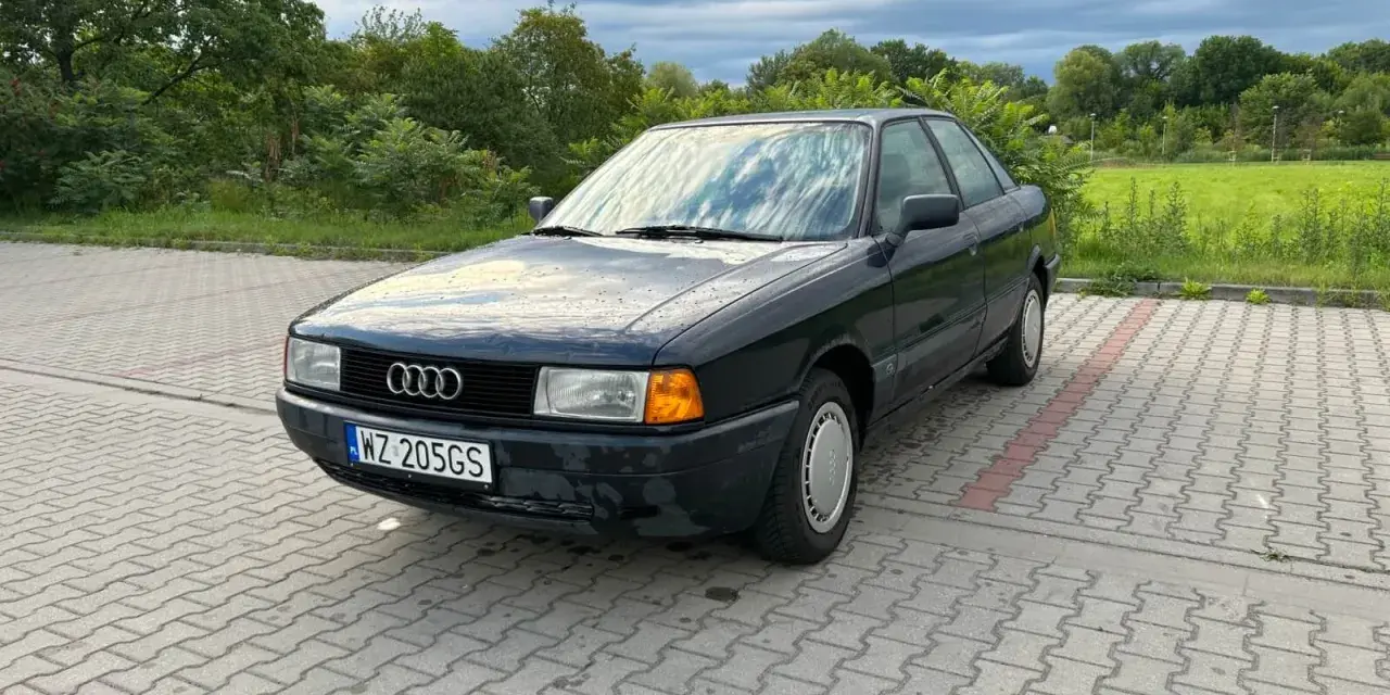 Ciemny Audi 80, klasyk z lat 90., idealny dla szukających aut do 5 tys. zł. Zaparkowany na kostce brukowej, otoczony zielenią.