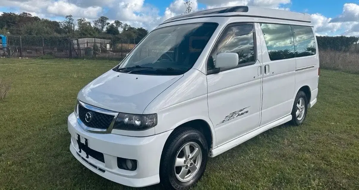 Biały van Mazda Bongo z podnoszonym dachem, zaparkowany na trawie.