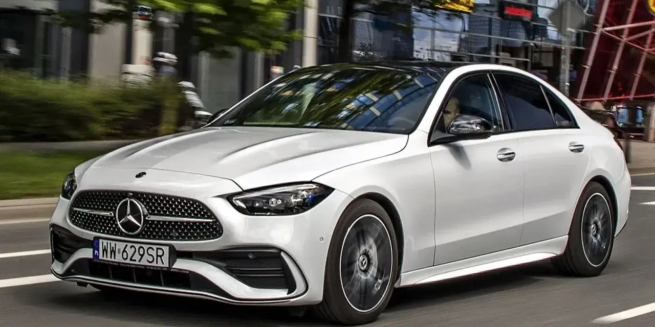 Biały Mercedes C 200 z czarnym dachem sunie po miejskiej drodze, odbijając światła budynków w szybach.