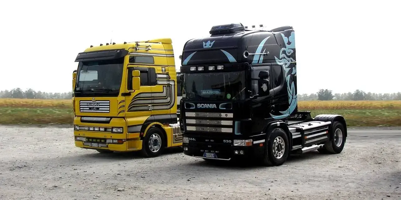 Dwa ciągniki siodłowe: żółty MAN i czarna Scania z grafiką. Różne rodzaje naczep czekają na załadunek.