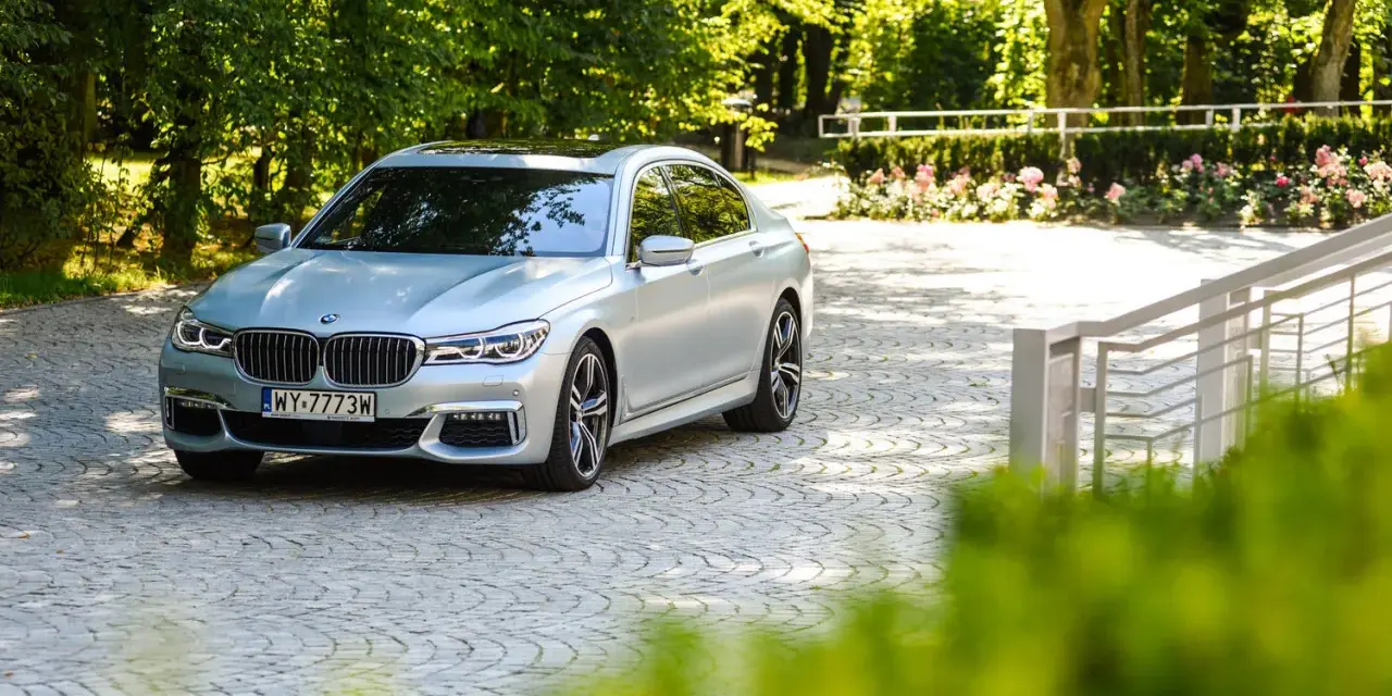 Srebrne BMW 750i lśni w słońcu na brukowanym podjeździe, otoczone zielenią i różami.