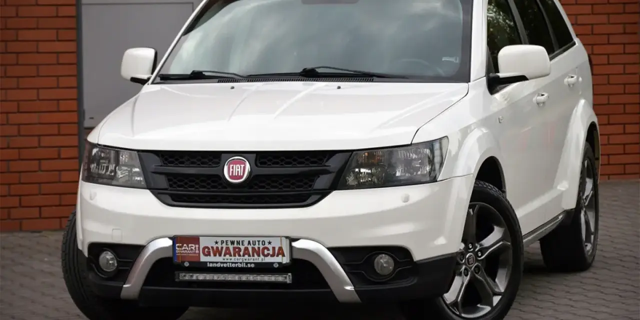 Biały Fiat 4x4 z czarną atrapą chłodnicy i chromowanymi elementami. Auto stoi na kostce brukowej przed budynkiem z cegły.