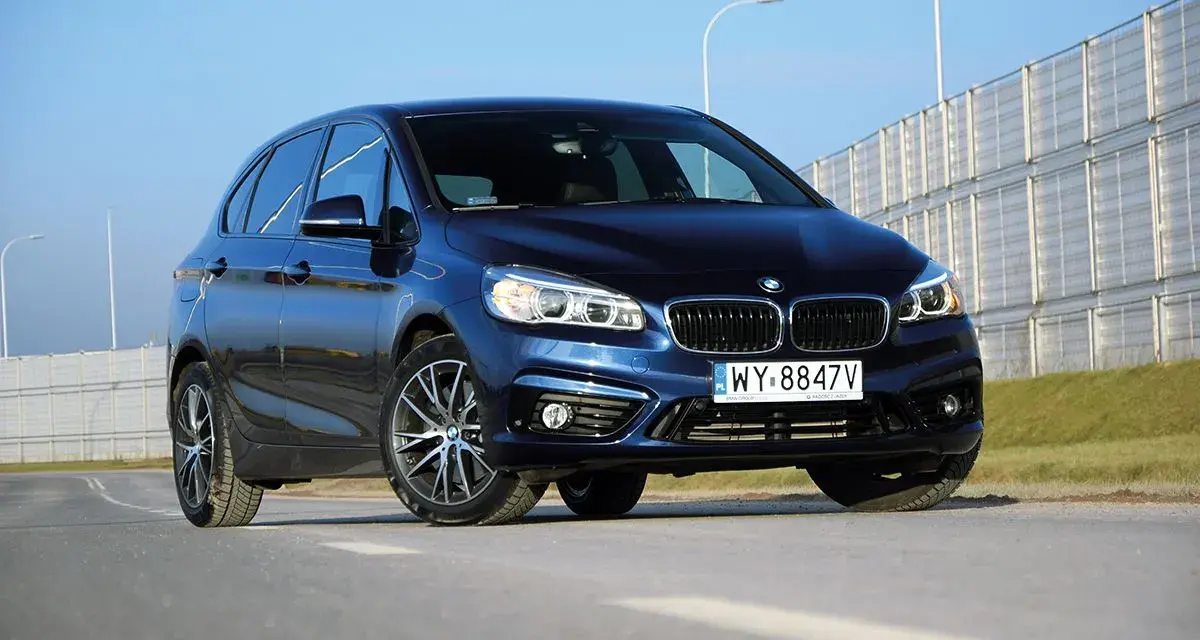 Ciemnoniebieski BMW van na tle jasnego nieba i barier dźwiękochłonnych.