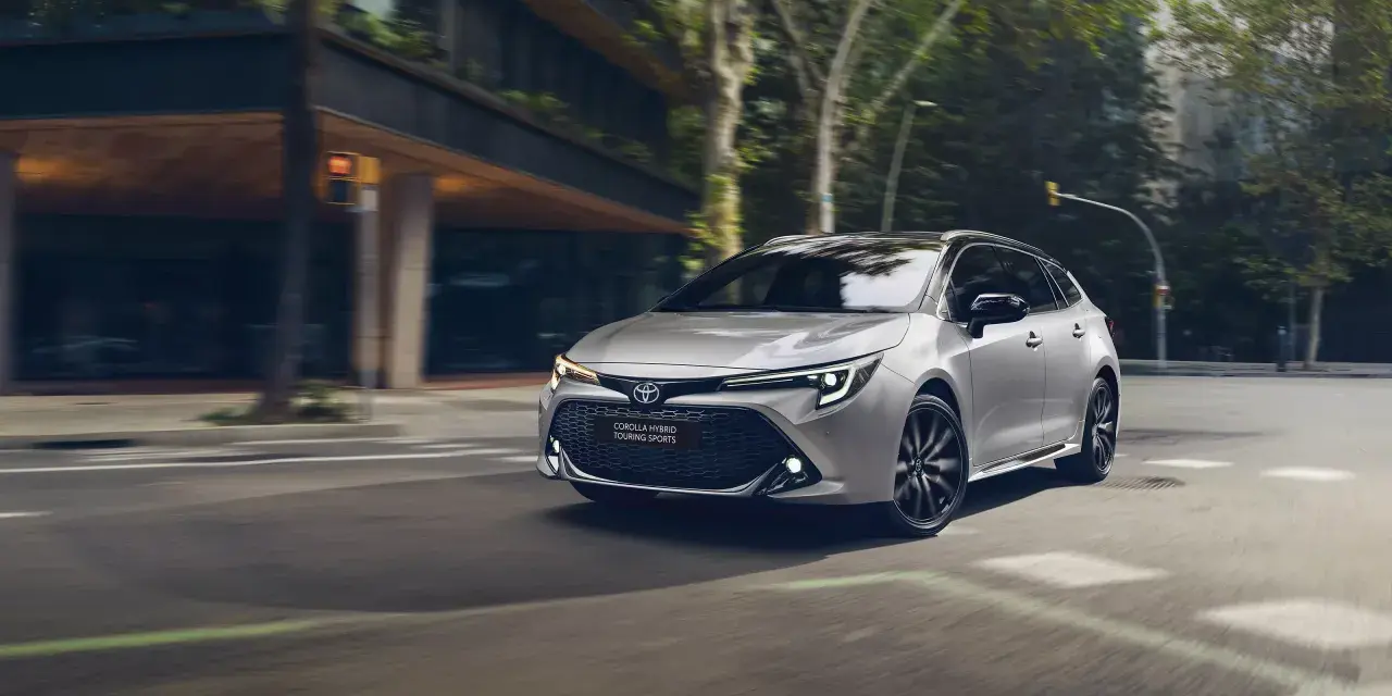 Srebrna Toyota Corolla kombi z czarnymi felgami sunie po miejskiej ulicy, otoczona zielenią i nowoczesną architekturą.