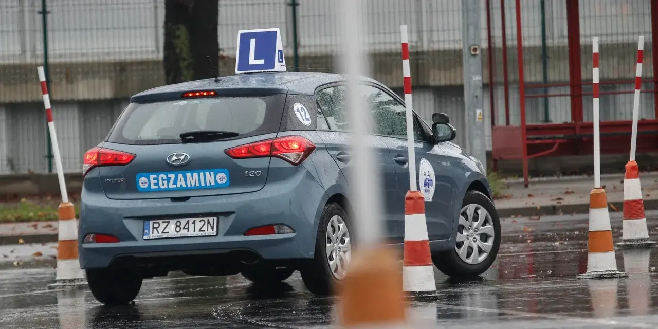 Samochód egzaminacyjny z literą "L" na dachu i napisem "EGZAMIN" na tylnej klapie, otoczony pachołkami na mokrym placu.