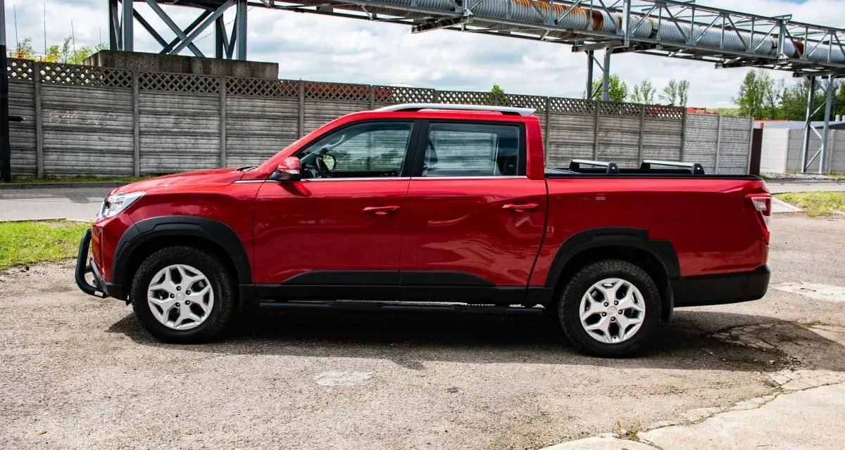 Czerwony SsangYong pickup z czarnymi dodatkami, zaparkowany na tle betonowego muru i przemysłowej konstrukcji.
