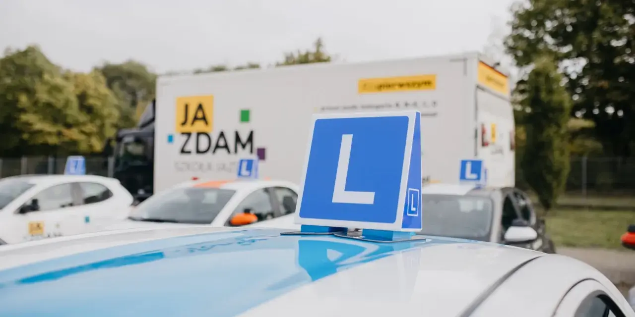 Na dachu samochodu nauki jazdy widoczny znak "L". W tle ciężarówka z napisem "JA ZDAM". Ile jest pytań na kat B?