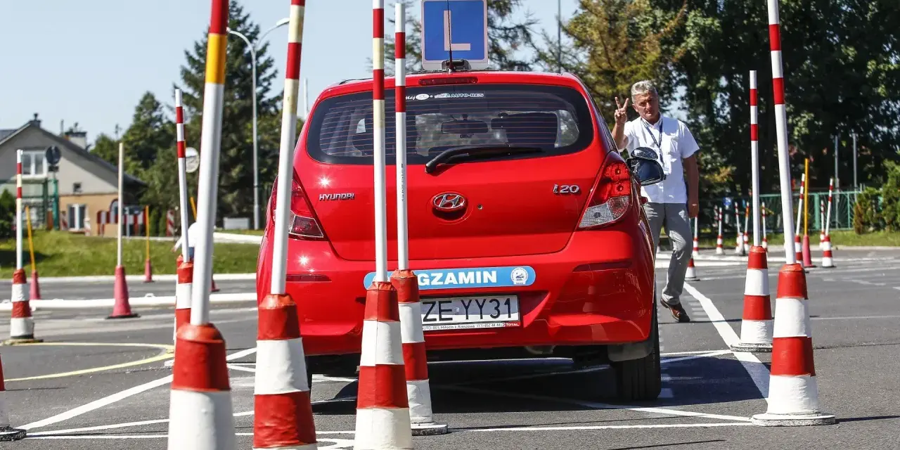 Czerwony Hyundai na placu manewrowym, z literą "L" na dachu. Instruktor pokazuje znak pokoju. Przygotowania do egzaminu na prawo jazdy b+e.