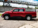 Czerwony SsangYong pickup z czarnymi dodatkami, zaparkowany na tle betonowego muru i przemysłowej konstrukcji.
