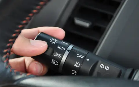 Dłoń ustawia przełącznik świateł w samochodzie, wybierając, na jakich światłach się jeździ: AUTO, OFF, światła mijania lub drogowe.