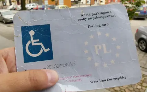 Karta parkingowa dla osoby z niepełnosprawnością, stopień umiarkowany. W tle samochody i budynki.
