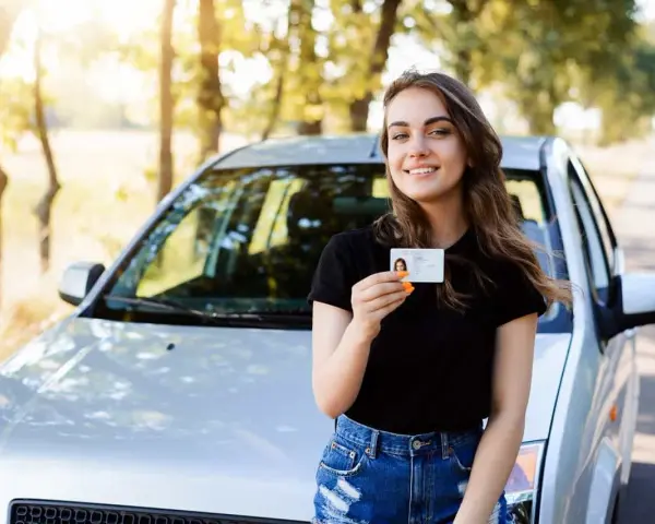 Szczęśliwa młoda kobieta z prawem jazdy od 16 lat stoi obok samochodu na tle drzew.