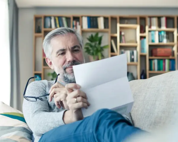 Mężczyzna z siwą brodą czyta dokument, być może wypowiedzenie OC. Na półkach za nim widać książki.
