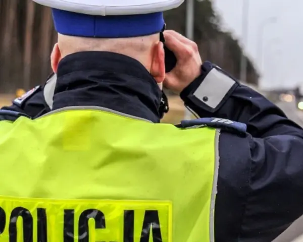 Policjant w kamizelce odblaskowej obserwuje drogę, gdzie widać ciężarówkę. Na drodze obowiązuje zakaz wyprzedzania dla ciężarówek.