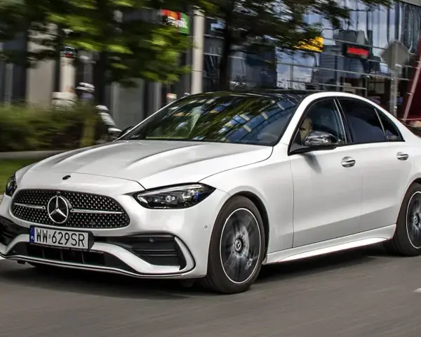 Biały Mercedes C 200 z czarnym dachem sunie po miejskiej drodze, odbijając światła budynków w szybach.
