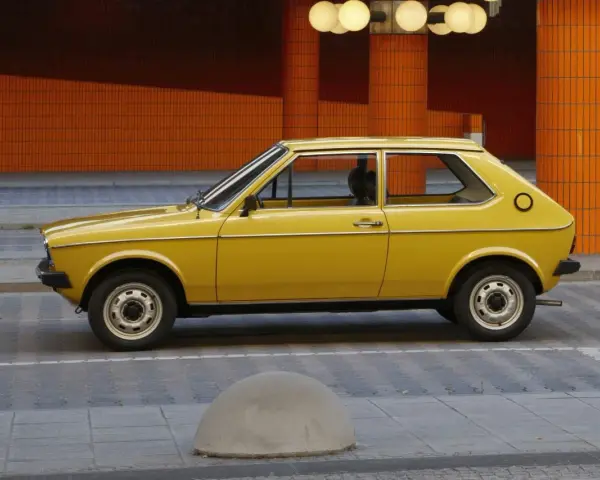Żółty Audi 50, klasyk z lat 70., parkuje na tle pomarańczowej, kafelkowej architektury.