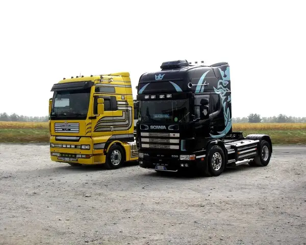 Dwa ciągniki siodłowe: żółty MAN i czarna Scania z grafiką. Różne rodzaje naczep czekają na załadunek.