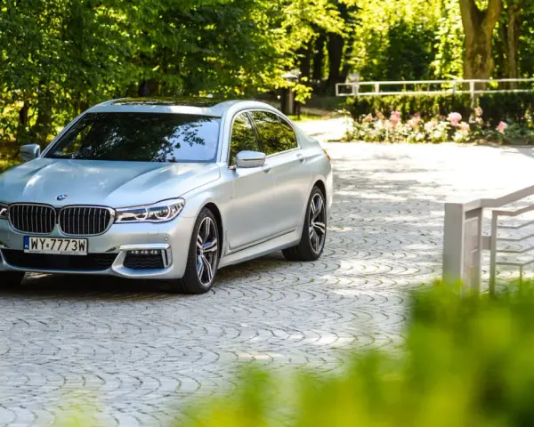 Srebrne BMW 750i lśni w słońcu na brukowanym podjeździe, otoczone zielenią i różami.