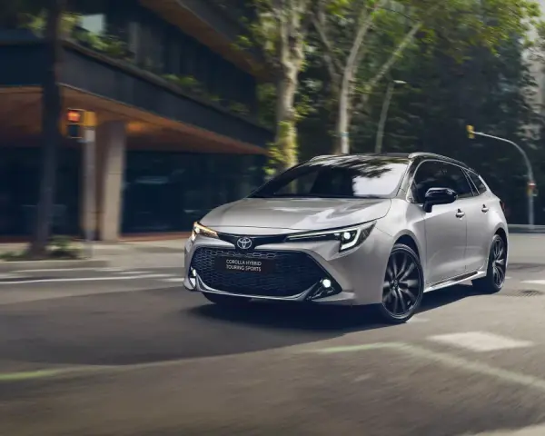 Srebrna Toyota Corolla kombi z czarnymi felgami sunie po miejskiej ulicy, otoczona zielenią i nowoczesną architekturą.