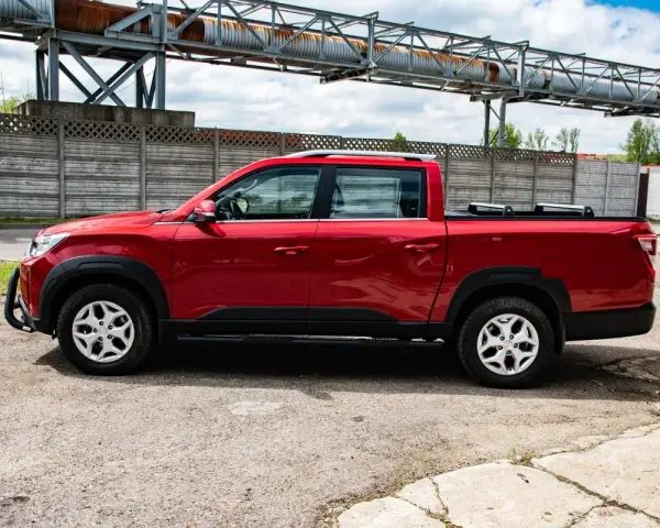 Czerwony SsangYong pickup z czarnymi dodatkami, zaparkowany na tle betonowego muru i przemysłowej konstrukcji.