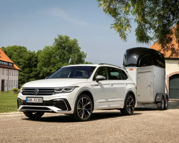 Biały Volkswagen SUV z przyczepą do przewozu koni stoi przed starym budynkiem.