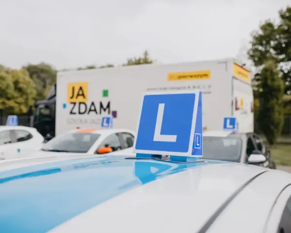 Na dachu samochodu nauki jazdy widoczny znak "L". W tle ciężarówka z napisem "JA ZDAM". Ile jest pytań na kat B?