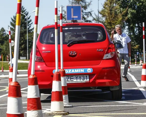 Czerwony Hyundai na placu manewrowym, z literą "L" na dachu. Instruktor pokazuje znak pokoju. Przygotowania do egzaminu na prawo jazdy b+e.