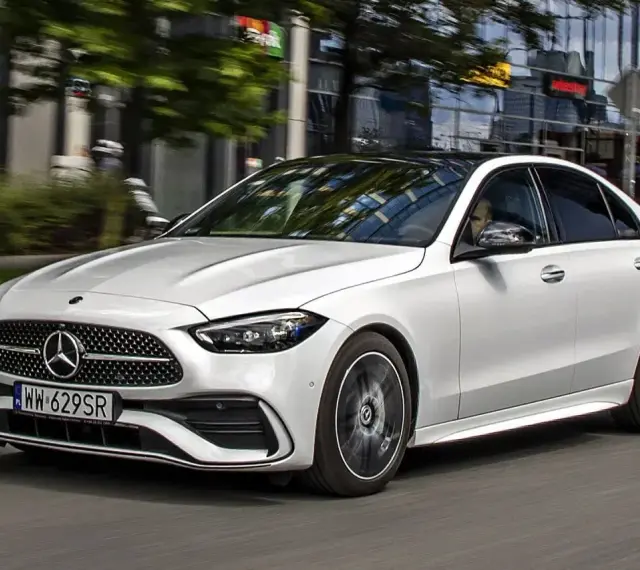 Biały Mercedes C 200 z czarnym dachem sunie po miejskiej drodze, odbijając światła budynków w szybach.