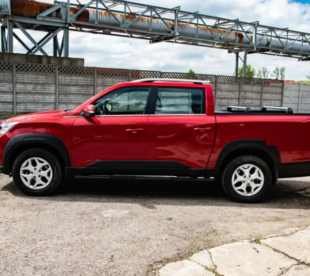 Czerwony SsangYong pickup z czarnymi dodatkami, zaparkowany na tle betonowego muru i przemysłowej konstrukcji.