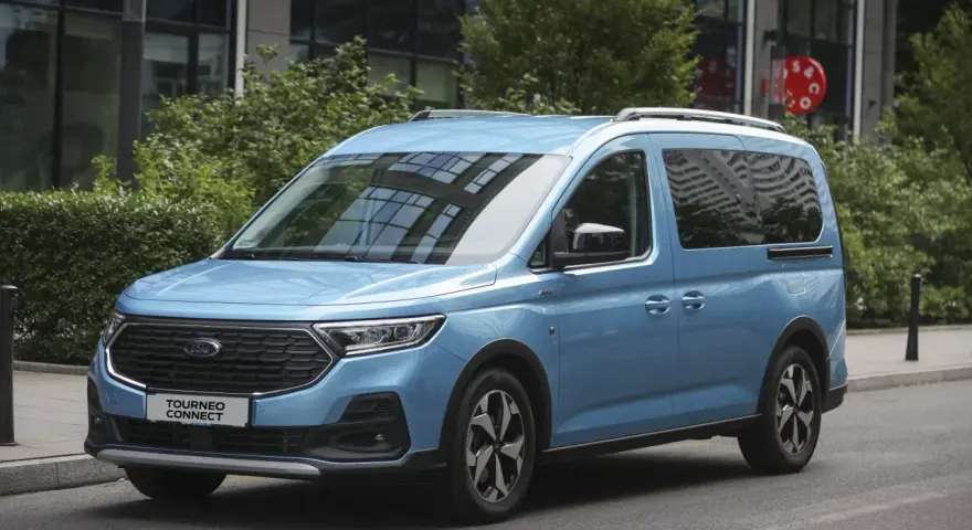 Niebieski Ford Tourneo Connect zaparkowany na ulicy, z budynkiem i zielenią w tle.
