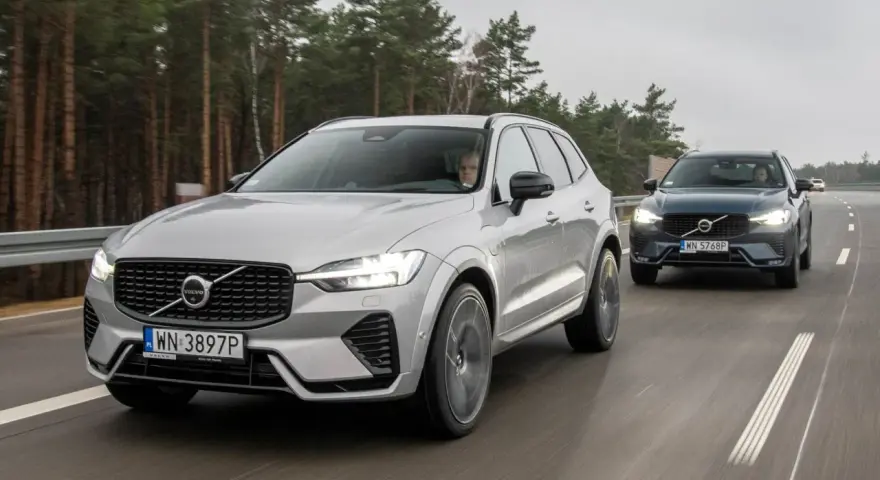 Dwa SUV-y Volvo jadące autostradą przez las. Srebrny SUV Volvo z przodu, ciemniejszy SUV Volvo za nim.