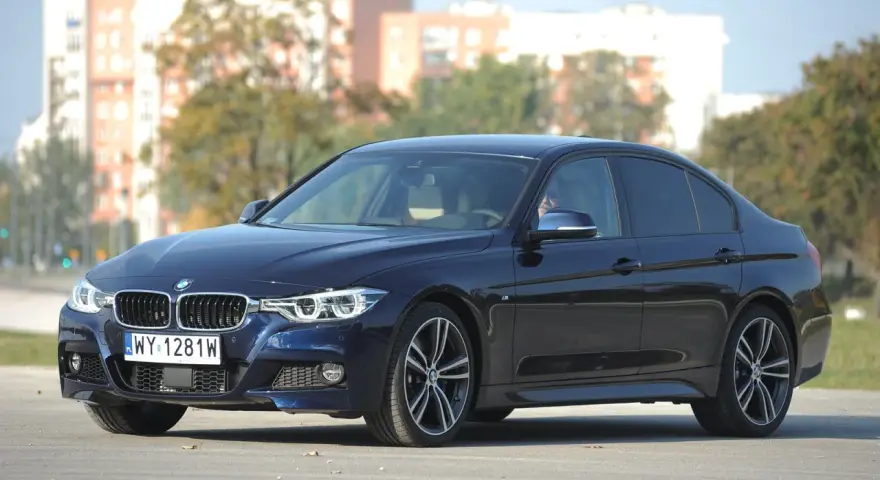 Ciemnoniebieskie BMW 318d z polskimi tablicami rejestracyjnymi stoi na drodze. W tle widać budynki i drzewa.