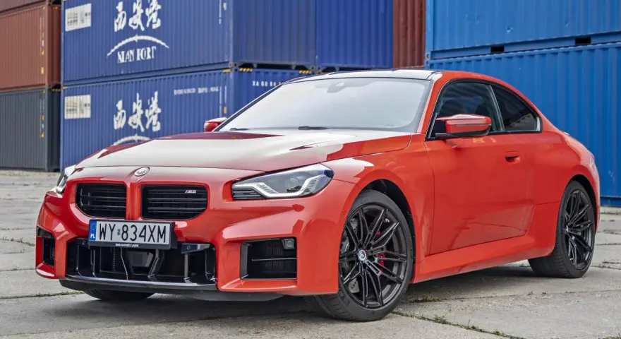 Pomarańczowe BMW M2 z czarnymi felgami, na tle niebieskich kontenerów. Sprawdź, jaka jest aktualna bmw m2 cena.