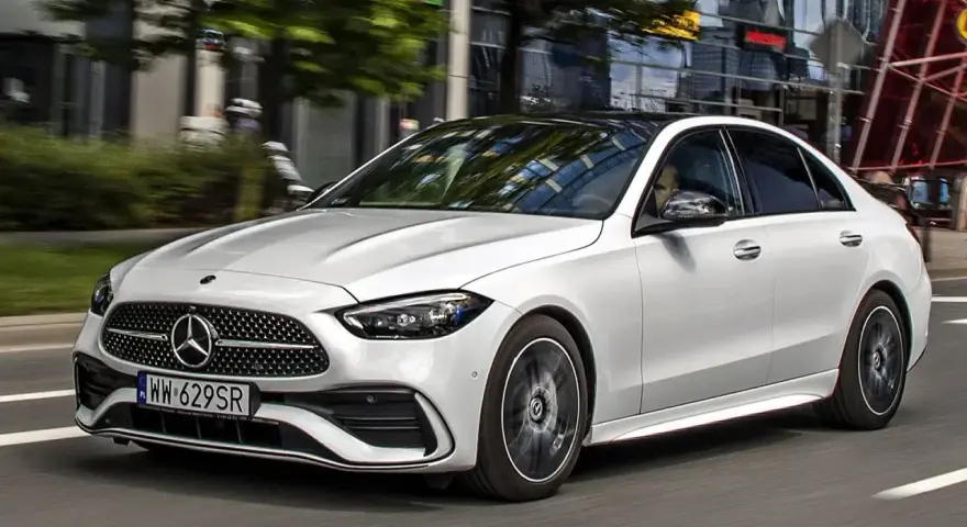 Biały Mercedes C 200 z czarnym dachem sunie po miejskiej drodze, odbijając światła budynków w szybach.