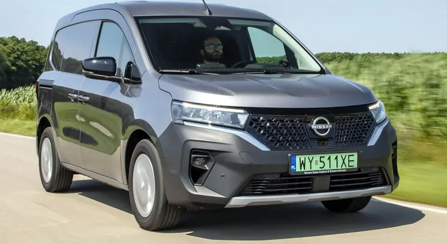Szary Nissan Townstar jedzie po drodze, otoczony zielonymi polami. Kierowca w okularach prowadzi pojazd.