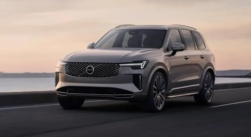 Elegancki, szary Volvo CX90 jedzie po drodze nad jeziorem o zachodzie słońca.