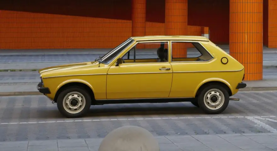 Żółty Audi 50, klasyk z lat 70., parkuje na tle pomarańczowej, kafelkowej architektury.