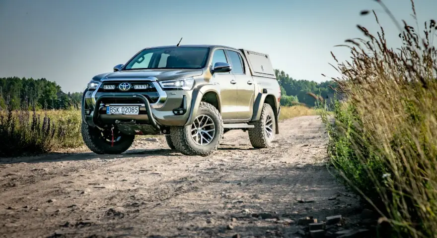 Złota toyota terenowa z wyciągarką i zabudową pędzi po polnej drodze, otoczona dziką roślinnością.